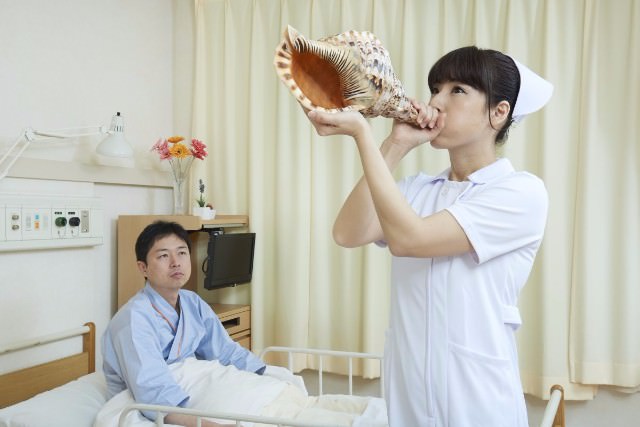 ベッドに座る治験被験者の横でほら貝を吹く看護師